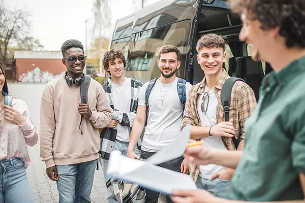 Diverse group of students getting on a minibus for a trip. The teacher is holding a clipboard and doing a headcount